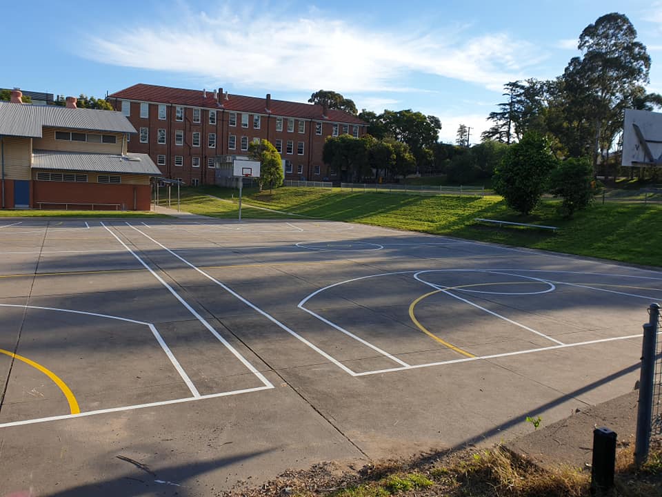 A basketball court with clear lines, behind a building and green trees on a sunny day.
